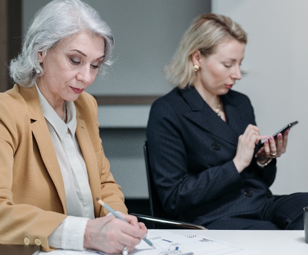 An elderly woman analysing a report next to a middle-aged woman working on a mobile device at a boardroom table in an office.